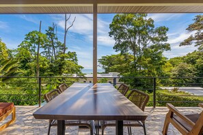 An outdoor dining area with a long table and chairs, surrounded by a railing, overlooking a lush landscape with trees and a view of the sea.