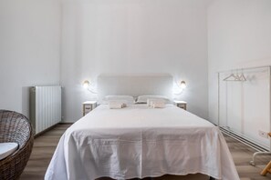 Une chambre à coucher avec un grand lit, des tables de chevet, un radiateur, un fauteuil en osier et un portant à vêtements.