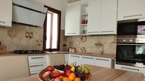 A modern kitchen with white cabinets, a gas stove, and a large oven. A fruit basket is on the counter. - Modern villa with garden in charming South Sardinia (Teulada)