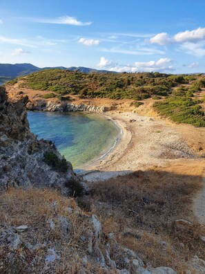 A coastal landscape with a clear beach, rocky outcrops, and a hilly backdrop.