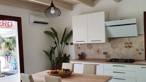 A modern kitchen with white cabinets, a wooden dining table, and chairs. There is a potted plant on the counter and a fruit bowl on the table. - Modern villa with garden in charming South Sardinia (Teulada)