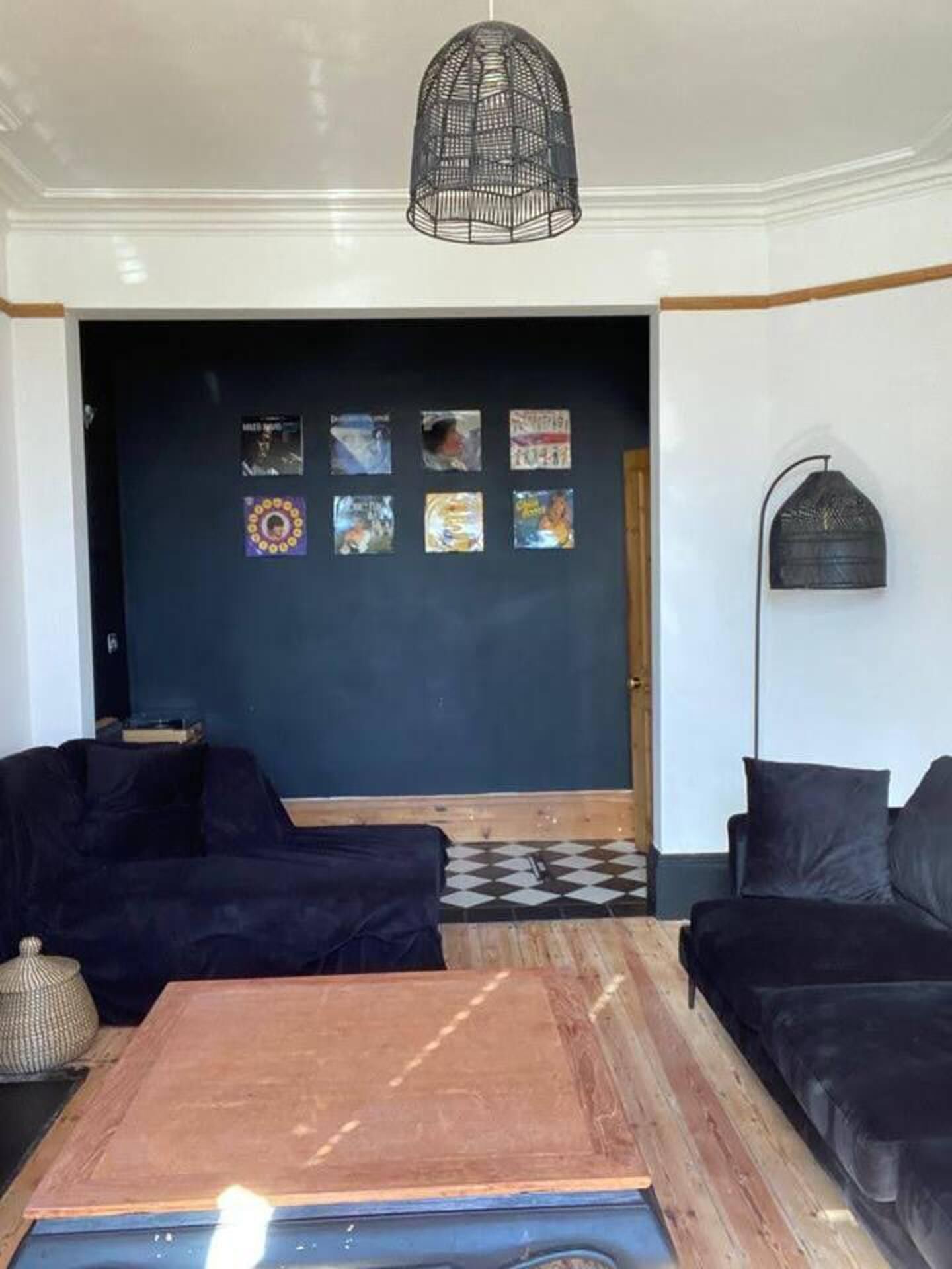 Un salon avec un mur bleu foncé, un luminaire suspendu en métal noir, deux canapés noirs, une table basse en bois et un lampadaire noir.