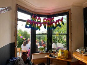 Quarto com uma janela grande, um banco de madeira, uma mesa com uma sacola e uma decoração de balão com a mensagem 'Happy Birthday'.