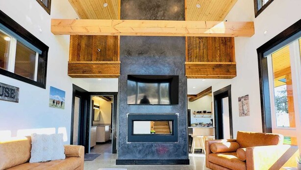 A modern living room with a fireplace, wooden ceiling, and large windows. - Eagle Cap Farms hosted owned and operated by Eagle Cap Vacation Rentals (Joseph)