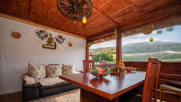 A wooden dining table with chairs, a bowl of fruit, and a view of a green landscape through a window.