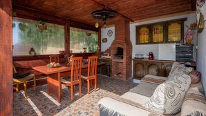 A rustic wooden interior with a stone-tiled floor, a dining area with a wooden table and chairs, a brick fireplace, and a sofa with patterned cushions.