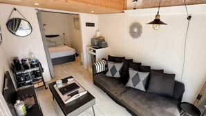 A modern living room with a dark brown sofa, a coffee table, and a small side table. There is a bed in the background, a mirror on the wall, and a shelf with various items. - Seaside Cabin Retreat in Hayling Island (Hayling Island)