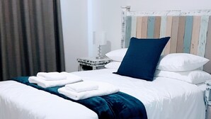 Una cama bien hecha con ropa de cama blanca y azul, una mesita de noche con una lámpara y un cuadro en la pared.