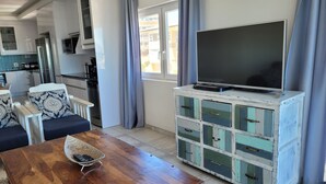 Una sala de estar moderna con una mesa de centro de madera, un televisor sobre un mueble blanco, y una cocina al fondo.