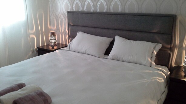 Een keurig opgemaakt bed met witte lakens en twee kussens, een nachtkastje met een lamp, en een behang met patroon.