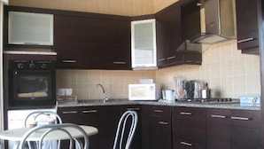 Een keuken met donkerbruine kastjes, een magnetron, een fornuis en een gootsteen. Er staan twee stoelen en een kleine tafel.