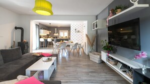 Een moderne woonkamer met een grijze bank, een witte salontafel en een televisie op een zwart dressoir. Achtergrond is een keuken met een eethoek.
