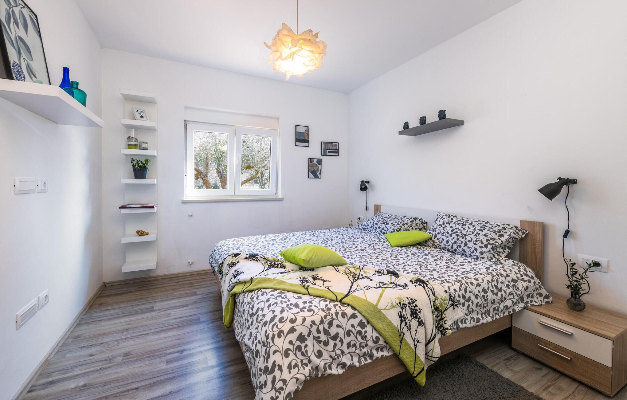 A bedroom with a bed, bedside table, lamp, shelf, window, and wall-mounted decor.