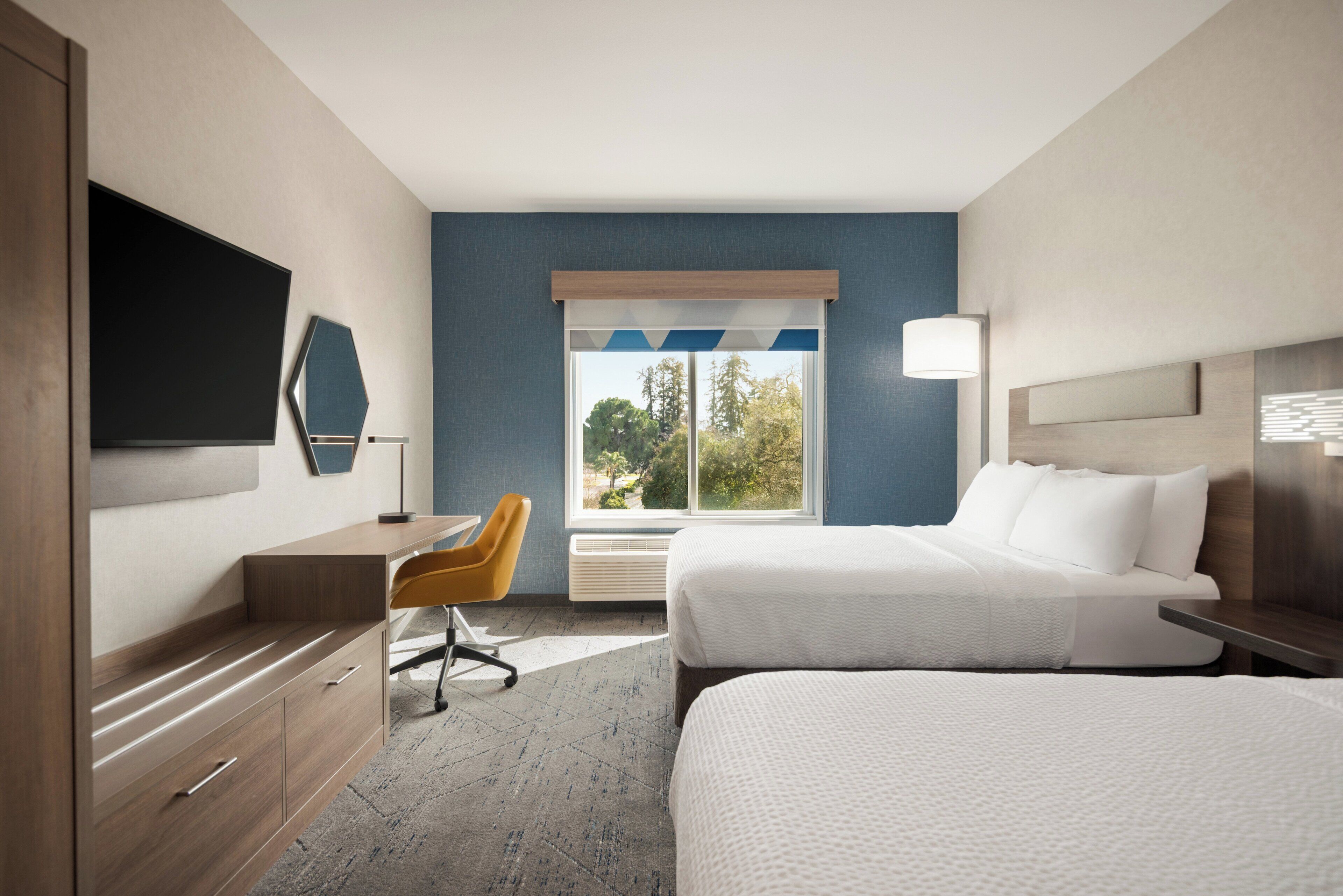 Habitación de hotel con cama, escritorio, silla, televisor y ventana con vistas a árboles.