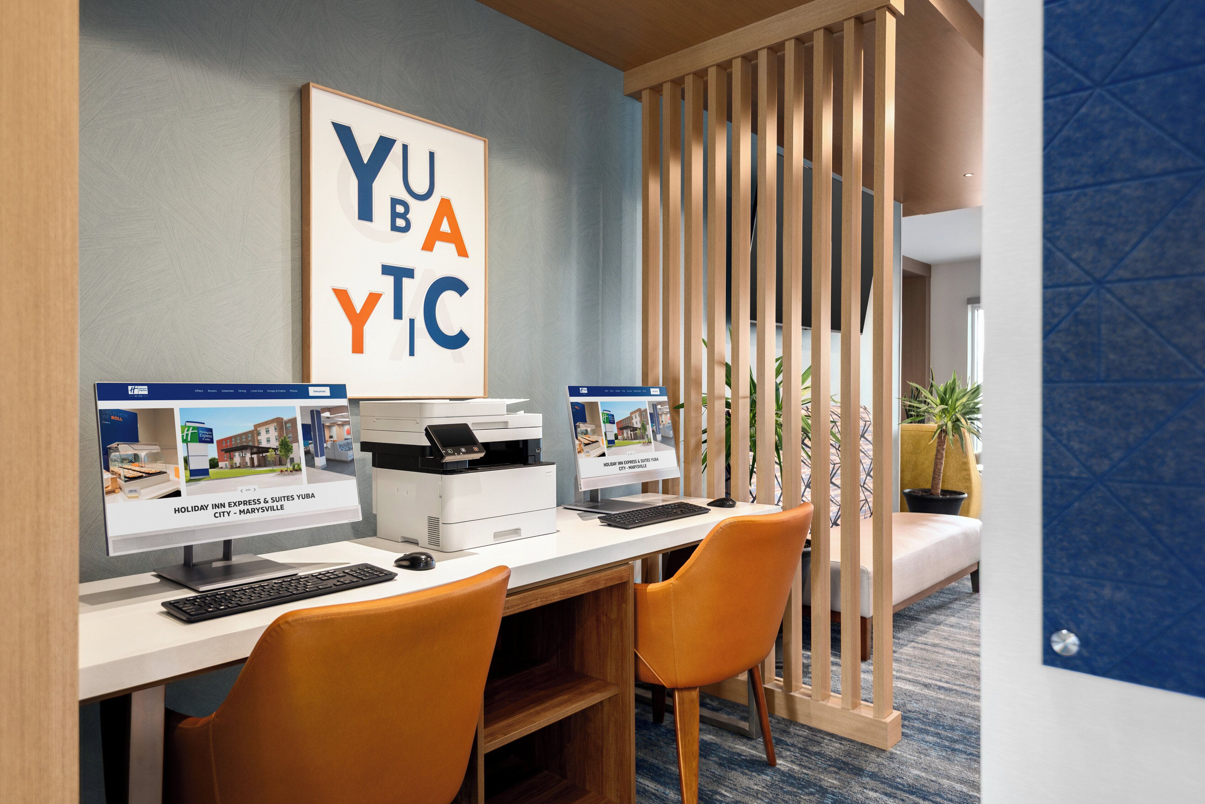 A hotel room with a desk, two monitors, a printer, and a large framed artwork.