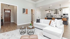 A spacious living room with a white sofa, a coffee table, and a rug. There is a kitchen in the background with modern appliances. - Luxe 3BR Family Stay | Fenced Yard | Prime WR (Kathleen)