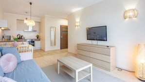 Ein modernes Wohnzimmer mit einer Couch, einem Couchtisch und einem Fernseher an der Wand. Im Hintergrund ist eine Küchenzeile mit einem Esstisch und Stühlen zu sehen.