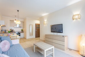 Ein modernes Wohnzimmer mit einer Couch, einem Couchtisch und einem Fernseher an der Wand. Im Hintergrund ist eine Küchenzeile mit einem Esstisch und Stühlen zu sehen.