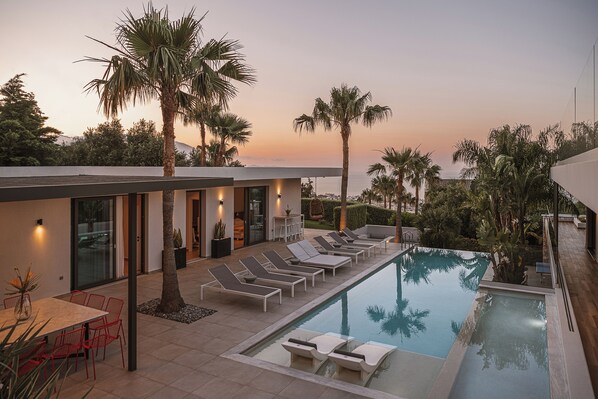 Un área de piscina moderna al aire libre con sillones de descanso, una mesa de comedor y vistas al atardecer.