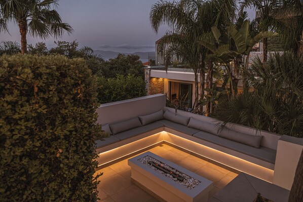 A modern outdoor seating area with a built-in fire feature, surrounded by lush greenery and overlooking a scenic landscape.