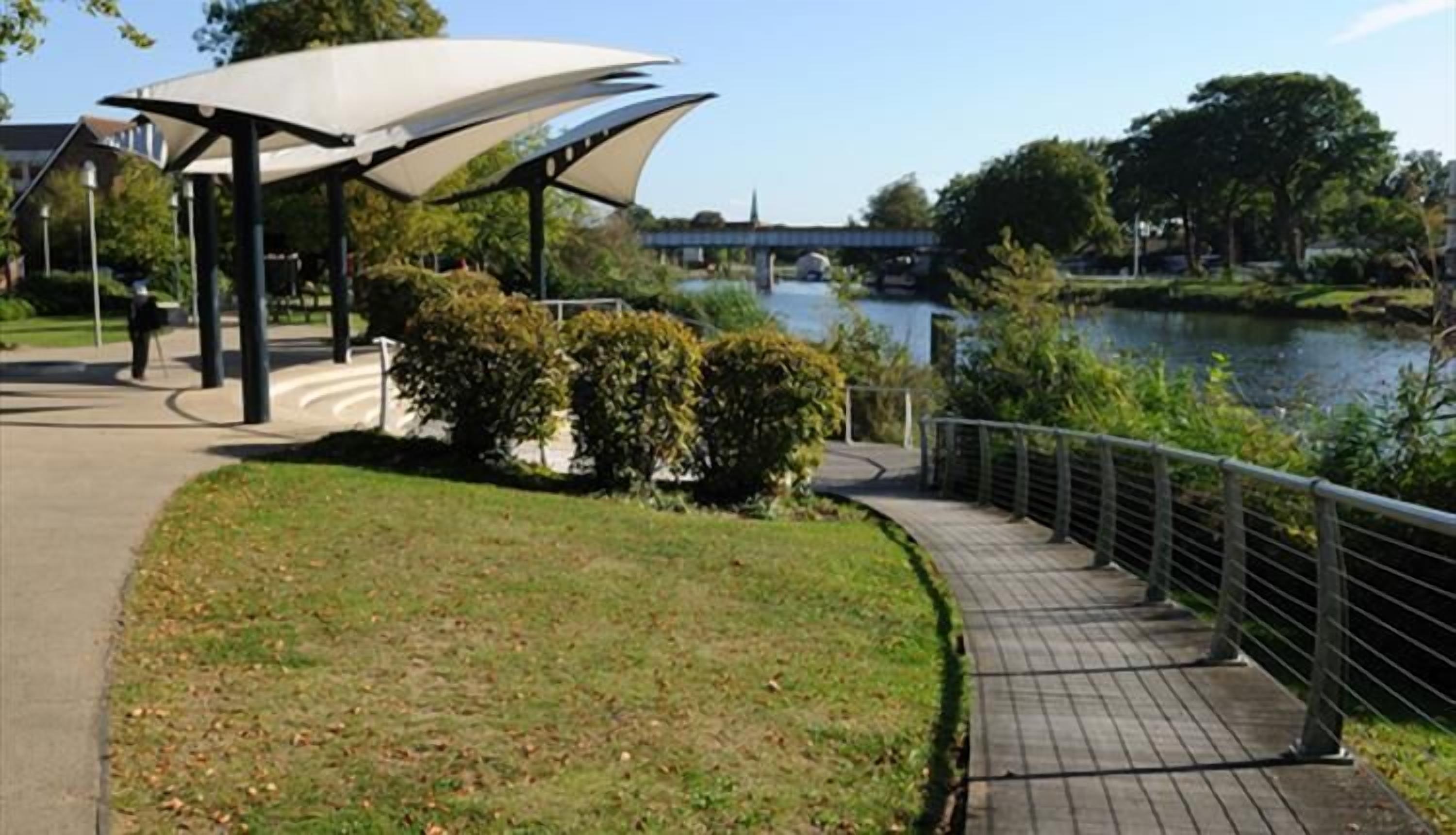 A park with a covered walkway, a river, and a bridge in the background.