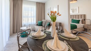 Een moderne hotelkamer met een eethoek voor vier personen, een bank, een bed met groene kussens en een groot raam met gordijnen.
