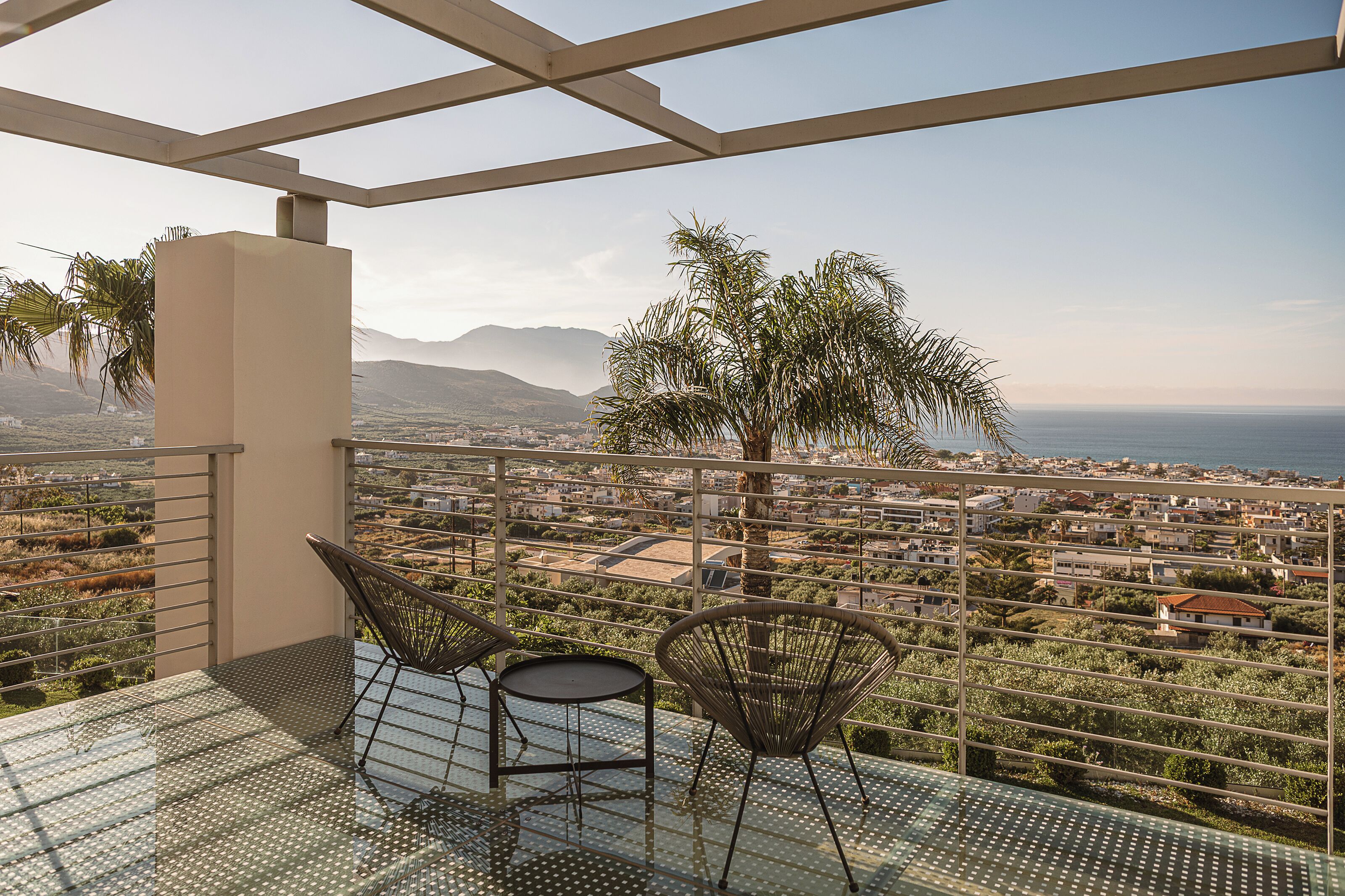 Ein Balkon mit Glastisch und Stühlen, der Blick auf eine Küstenstadt mit Bergen in der Ferne gewährt.