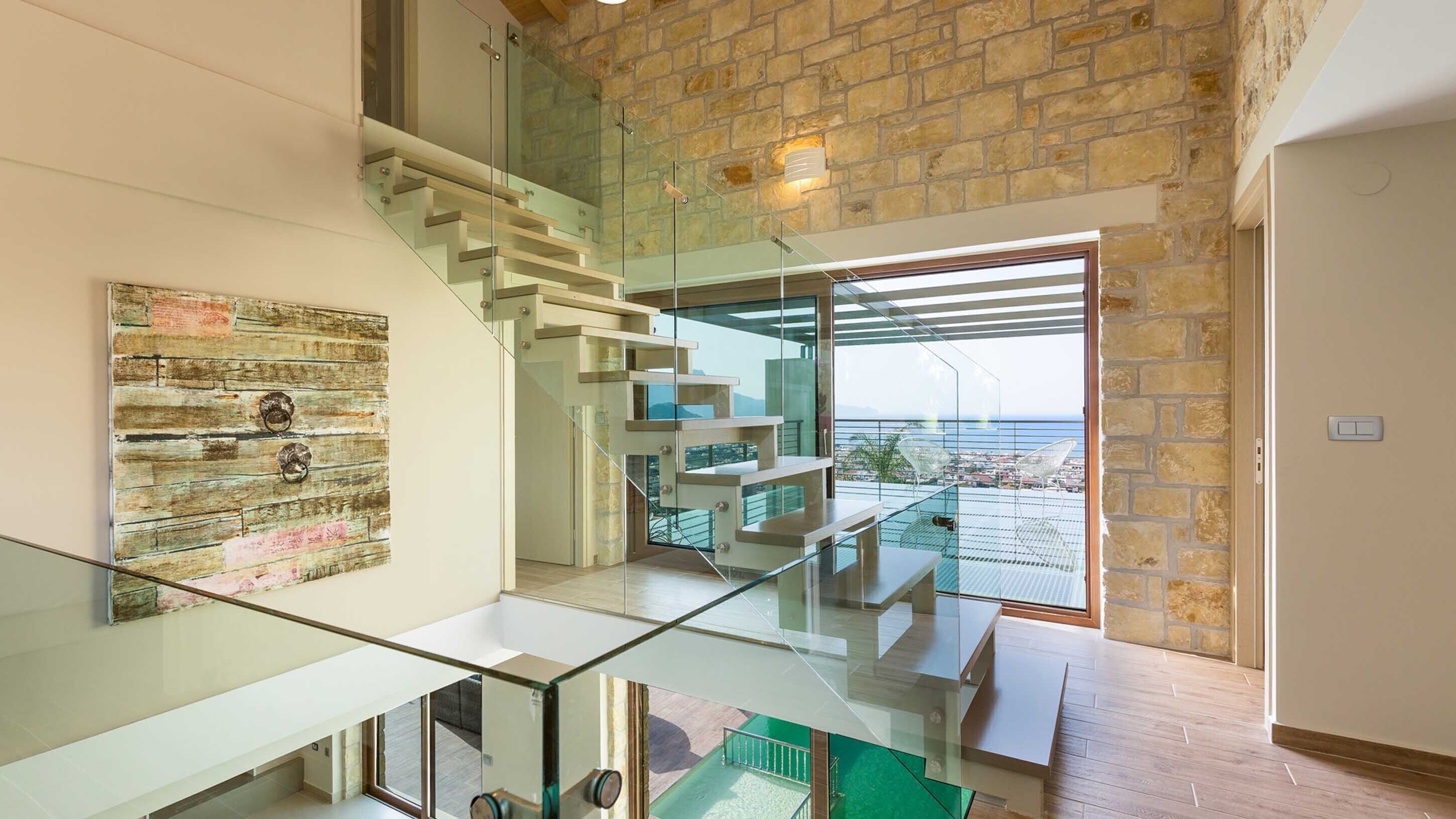 A modern interior with a glass staircase, a view of the sea, and a rustic wall.