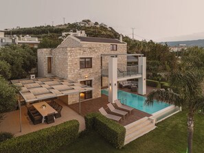 Une maison en pierre à deux étages, dotée d’une terrasse sur le toit, d’une piscine et d’un patio aménagé avec du mobilier d’extérieur.