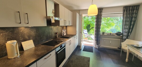 A modern kitchen with a black countertop, white cabinets, and a built-in oven. There is a window with curtains, a radiator, and a view of greenery outside.