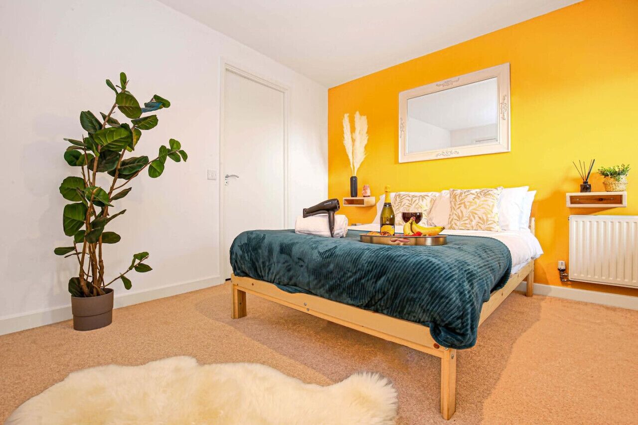 Ein Schlafzimmer mit einem Bett, einer Zimmerpflanze, einem Spiegel und einem Nachttisch mit Lampe und Flasche.