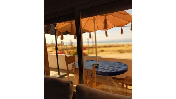 Ein Zimmer mit großem Fenster mit Blick auf einen Strand, ein Tisch mit blauer Tischdecke und ein Stuhl.