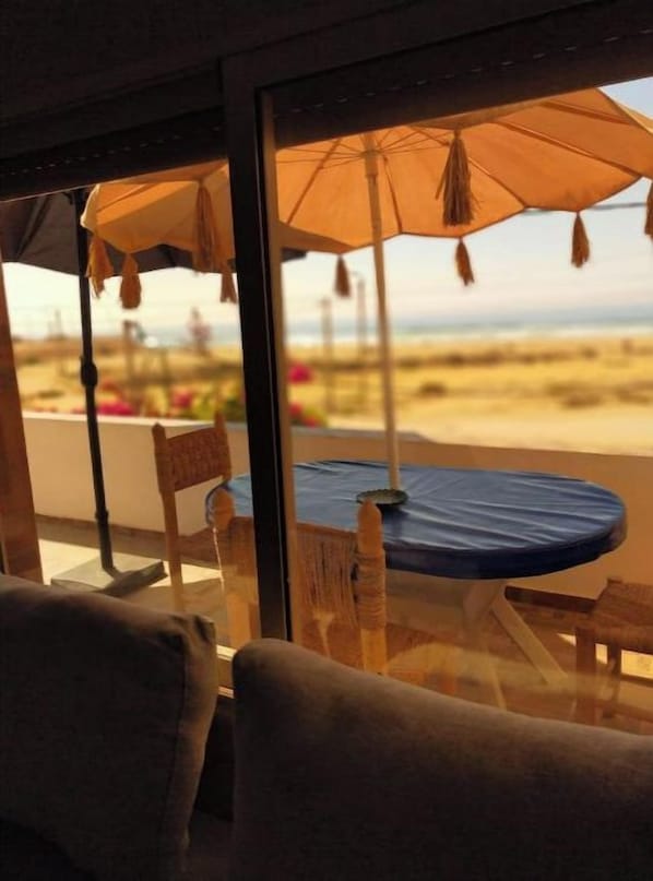 A room with a large window overlooking a beach, a table with a blue tablecloth, and a chair.