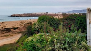 Eine Küstenlandschaft mit Sandstrand, Felsen und einem Gebäude im Hintergrund.