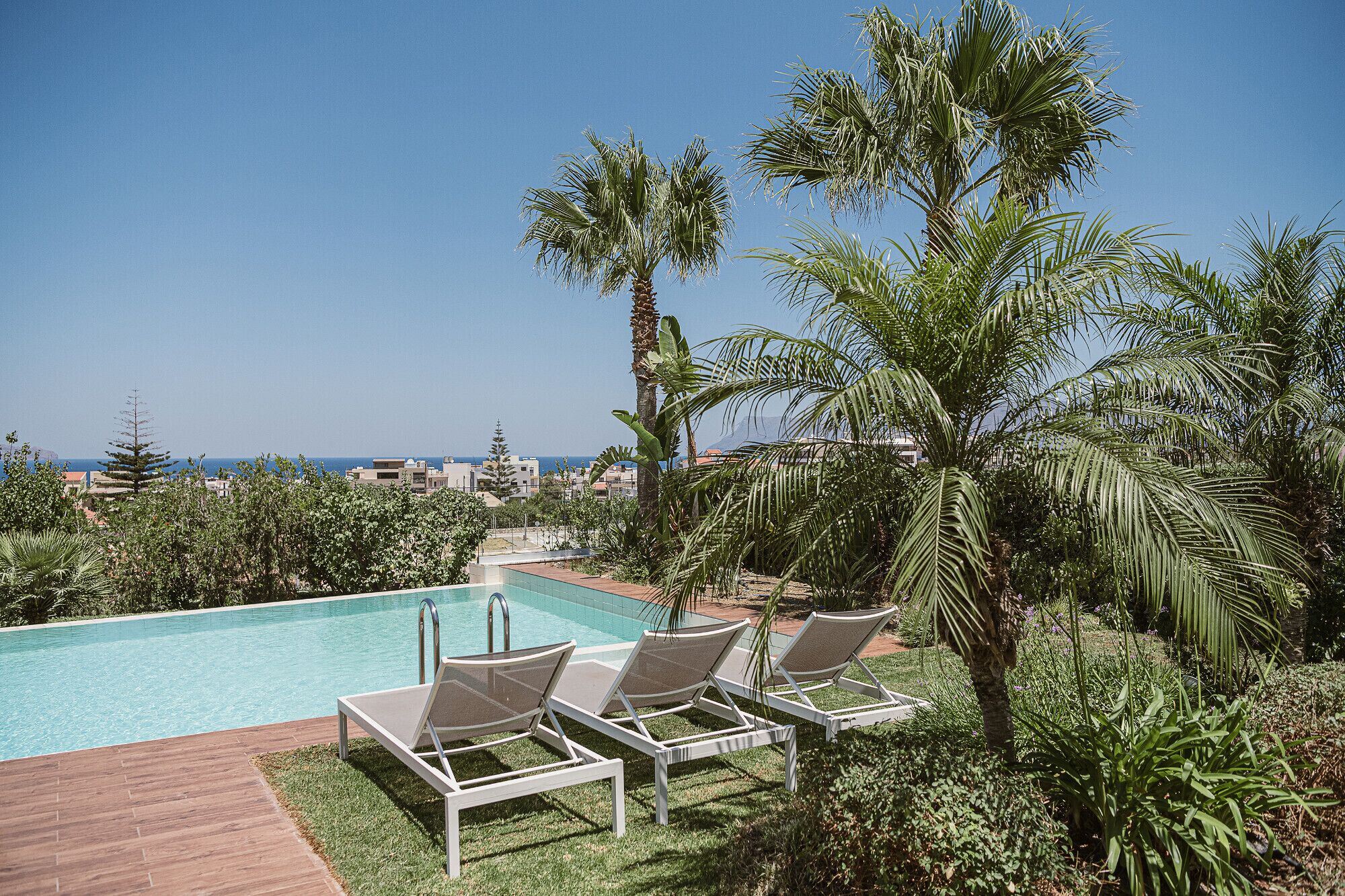Zona de piscina con sillones de descanso, palmeras y vista al mar.