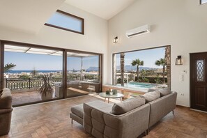 Ampla sala de estar com um sofá grande, uma mesa redonda pequena e vista para uma piscina e paisagem urbana.