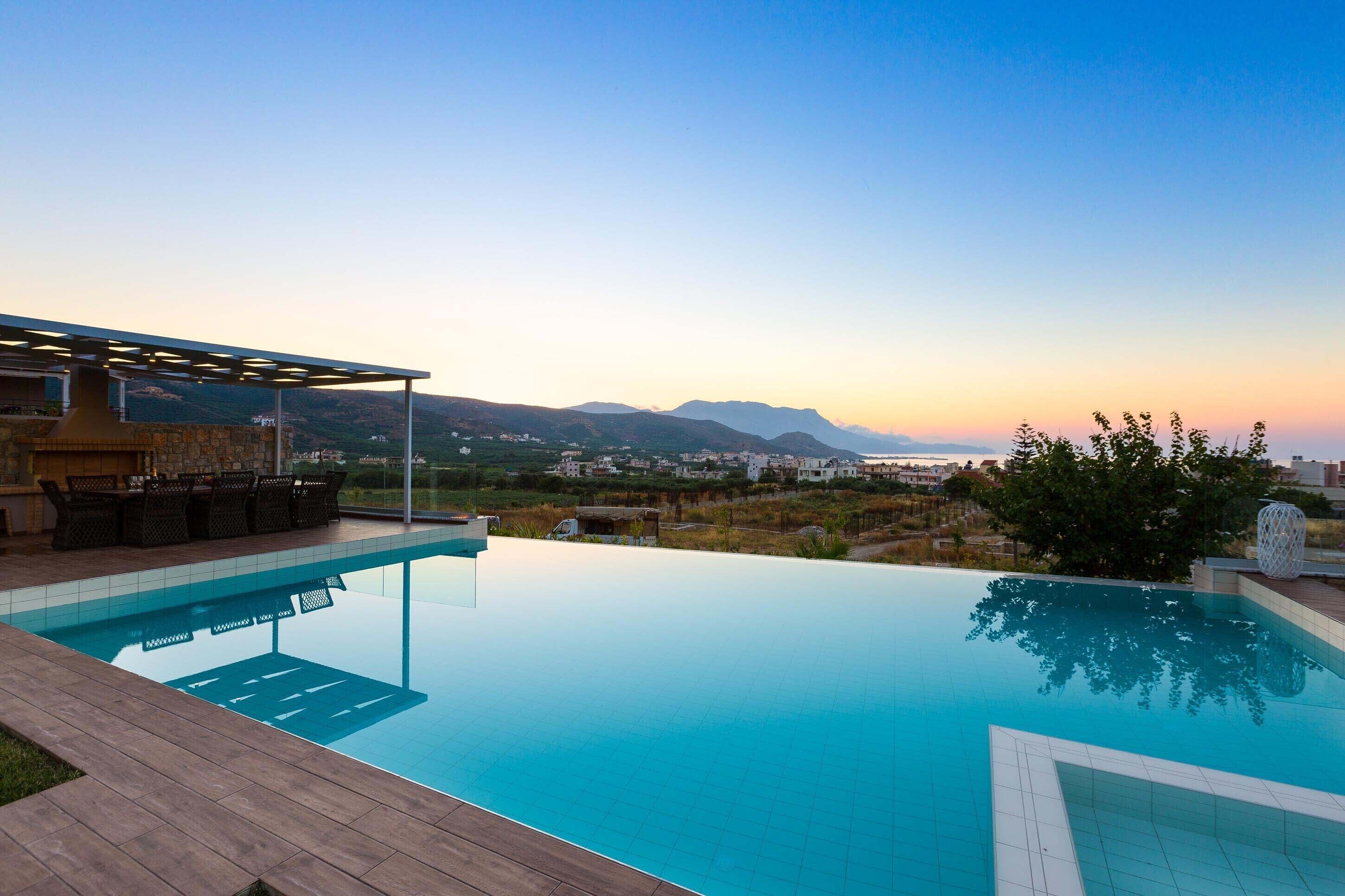 Una piscina infinita con vistas a montañas distantes y un patio con mobiliario exterior.