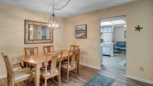 A dining area with a wooden table and chairs, a chandelier, a framed mirror, and a painting on the wall. - Coastal Oaks - Atlantic Beach home just 3 blocks from the beach (Atlantic Beach)