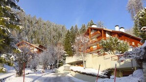 Ein schneebedecktes Gebäude mit Balkon, umgeben von Bäumen und bewölktem Himmel.