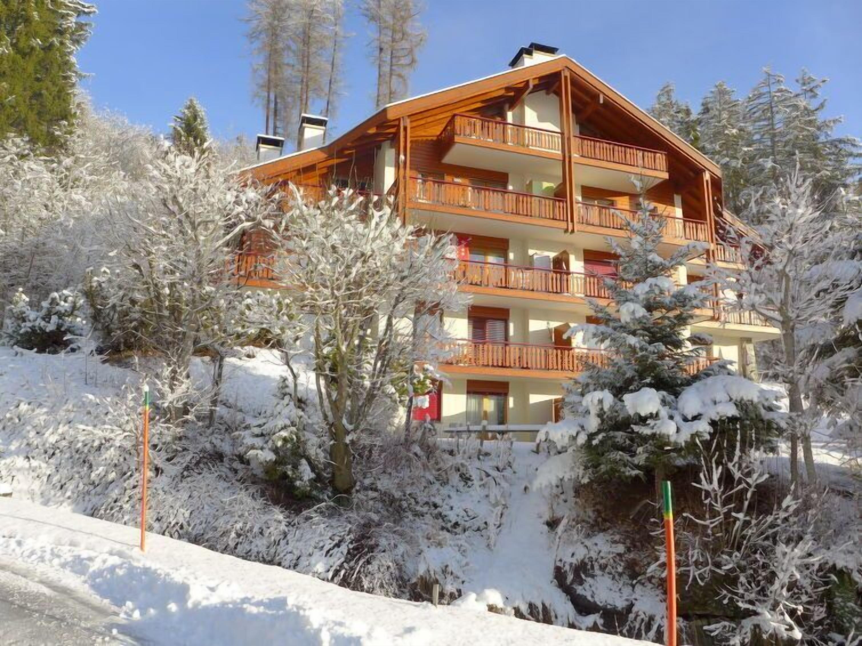 A multi-story wooden building with balconies, surrounded by snow-covered trees and a clear sky.