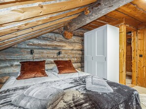 Ein Schlafzimmer mit schrägem Dach, Wänden aus Holzbalken, einem Bett mit gemustertem Überwurf, zwei braunen Kissen und einem weißen Kleiderschrank.