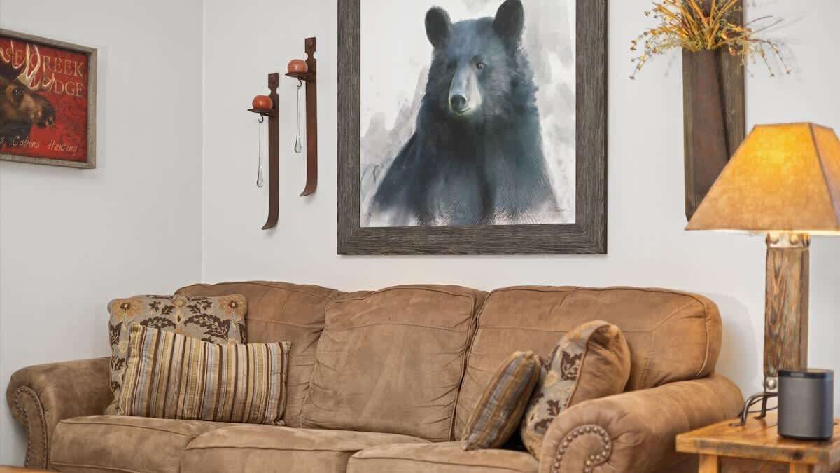 Un canapé en cuir marron agrémenté de coussins à motifs, un tableau représentant un ours encadré, une horloge murale rustique et une lampe posée sur une table d’appoint en bois.