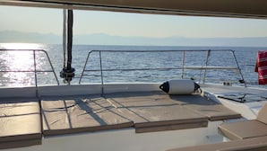 Een scheepdek met zitgedeeltes met kussens, een stuurwiel en uitzicht op de zee en verre bergen.
