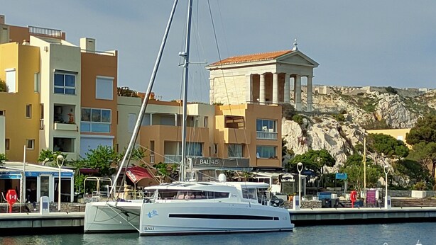 Een jachthaven met een aangemeerd jacht, gebouwen op de achtergrond en een rotsachtige heuvel.