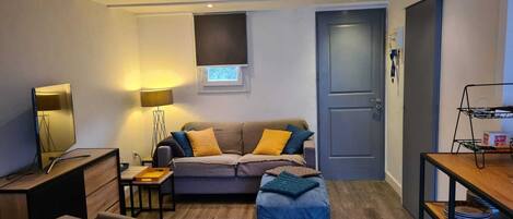 Un salon moderne avec un canapé, un pouf et une table à manger. On y trouve une lampe, une petite table avec un plateau et une étagère avec des assiettes.