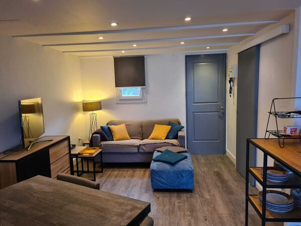 A modern living room with a sofa, ottoman, and a dining table. There is a lamp, a small table with a tray, and a shelf with plates. - Beautiful duplex apartment for 4 people in Tiuccia (Casaglione)