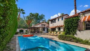 A swimming pool with lounge chairs and a patio area, surrounded by greenery and buildings with Mediterranean architecture.