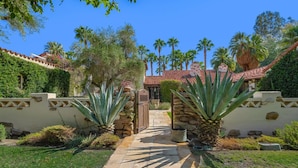 A stone pathway leading to a building with a red-tiled roof, surrounded by palm trees and a well-maintained garden.