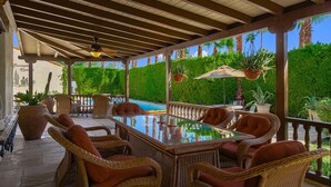 A covered outdoor seating area with wicker furniture and a glass table, overlooking a pool and lush greenery.