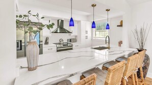 A modern kitchen with white cabinetry, a central island, and blue pendant lights. - Anna Maria Gem! Pool, Canal Views and Style! (Anna Maria)